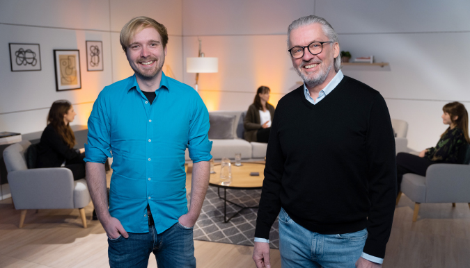 Stefan Grimm und Gerd Wutzler in einem stilvollem Studio, im Hintergrund finden Gespräche in einer gemütlichen Lounge-Atmosphäre statt.