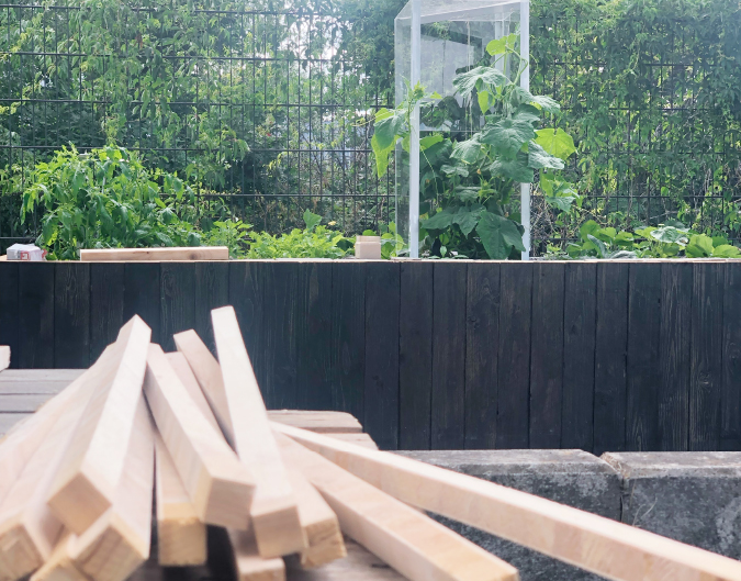 Stapel von Holzleisten im Vordergrund, dunkles Hochbeet mit Pflanzen und einem transparenten Schutzgehäuse für eine Pflanze im Hintergrund.