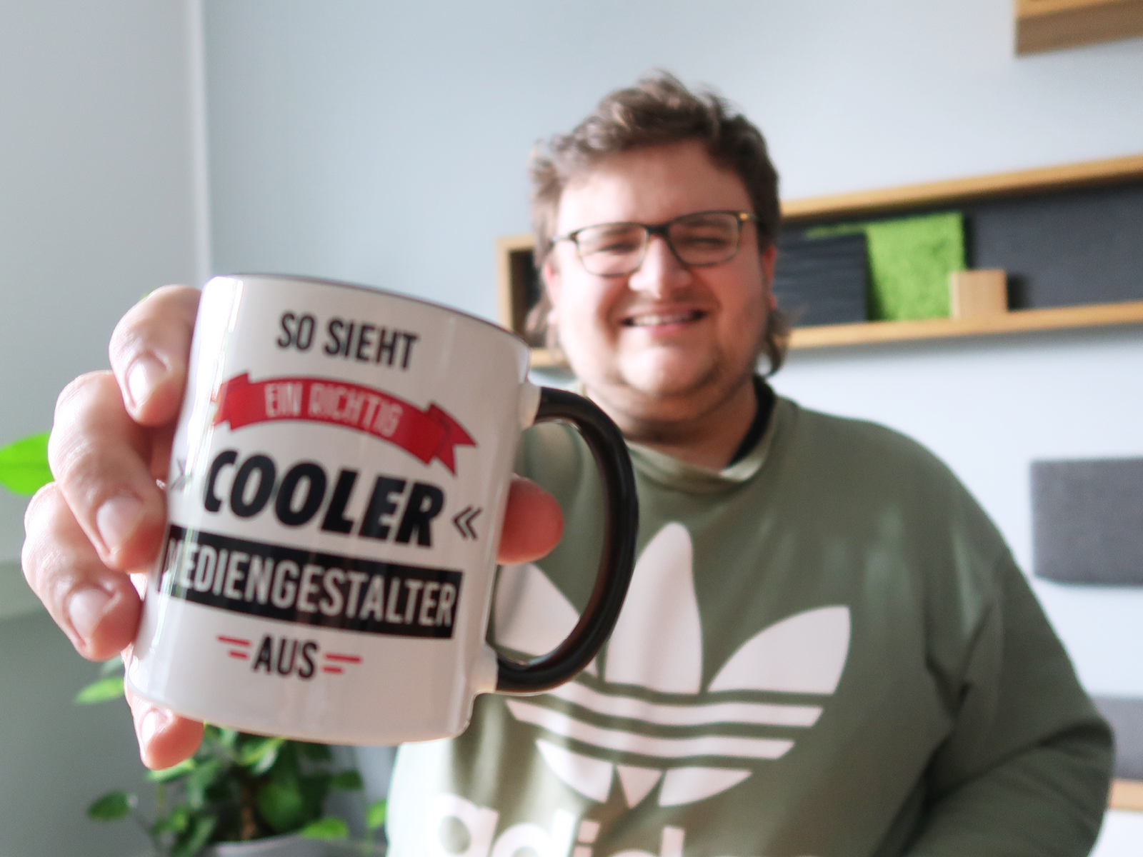 Ein lächelnder junger Mann mit Brille und grünem Adidas-Hoodie hält eine weiße Tasse mit schwarzem Henkel in die Kamera. Auf der Tasse steht: „So sieht ein richtig cooler Mediengestalter aus“. Im Hintergrund ein modernes Büro mit Pflanzen und Wandregalen.