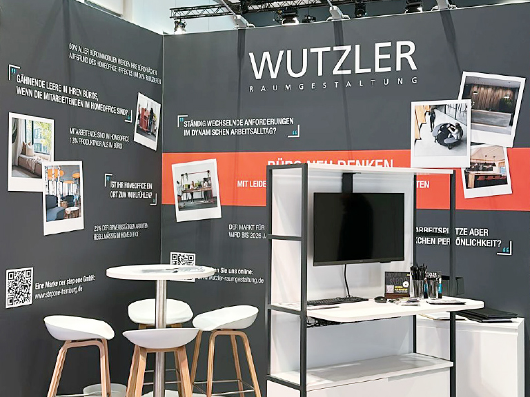 Messestand von Wutzler Raumgestaltung mit einem Bildschirm auf einem Regal, runden Stehtischen mit weißen Hockern und einer Rückwand voller inspirierender Fragen und Bildmotiven zu modernen Arbeitswelten.