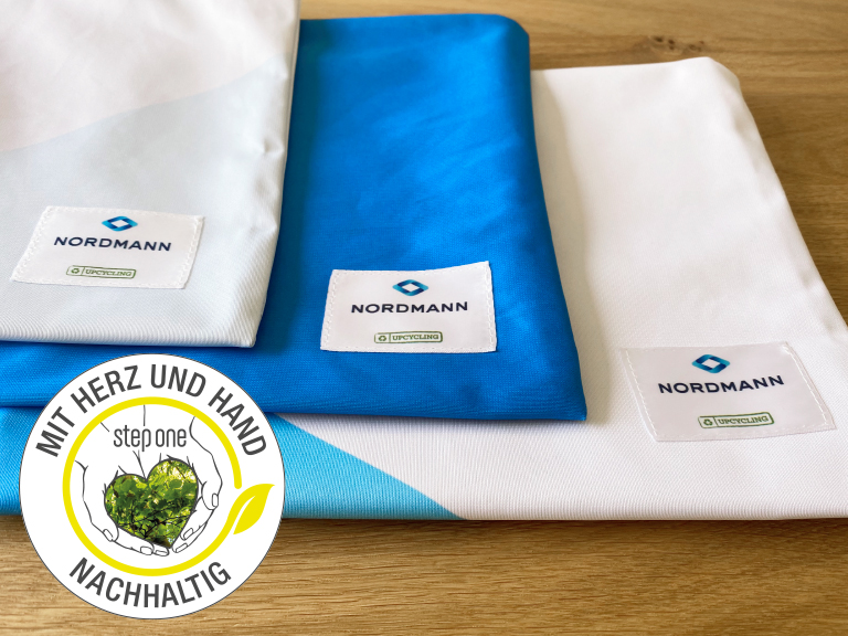 Drei farbige Stofftaschen mit Nordmann-Logo und Upcycling-Label auf Holztisch; unten links ein rundes Nachhaltigkeitssiegel mit Herzmotiv und der Aufschrift „Mit Herz und Hand – nachhaltig“.