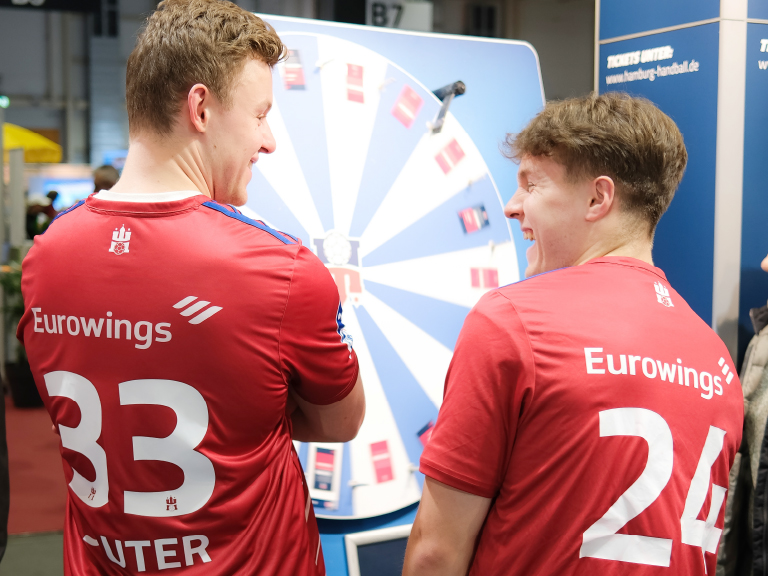 Zwei HSV-Handballspieler im roten Trikot stehen lachend vor einem großen Glücksrad an einem Messestand.