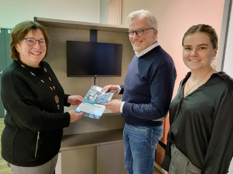 Zwei Frauen und ein Mann stehen lächelnd nebeneinander. Zwei von ihnen halten gemeinsam ein Projektblatt vor einem Möbelstück mit integriertem Bildschirm. Alle wirken freundlich und engagiert.