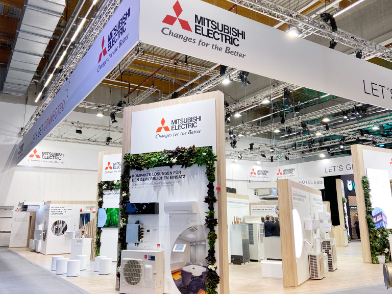 Großer Messeauftritt von Mitsubishi Electric. Der Stand zeigt verschiedene Klimageräte und technische Lösungen, umrahmt von Pflanzen und hellem Holzdesign. Über dem Stand hängt ein weißes Banner mit dem Firmenlogo.