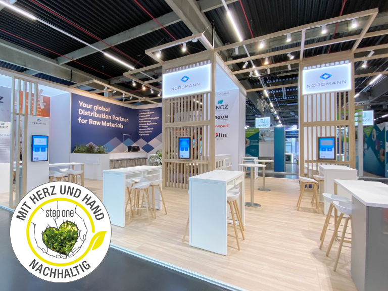 Heller Messestand von Nordmann mit Holz-Elementen, weißen Sitzmöbeln, digitalen Bildschirmen und einem Nachhaltigkeitssiegel – eine moderne und einladende Atmosphäre.