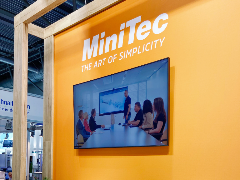 Orangene Messewand mit großem Monitor zeigt MiniTec-Präsentation; darüber der Slogan „MiniTec – The Art of Simplicity“.