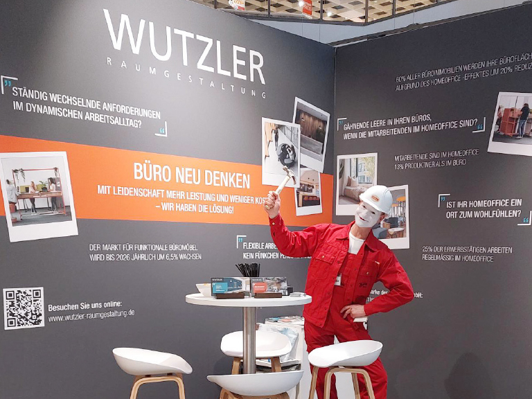 Ein Pantomime in rotem Monteuranzug mit weißem Helm steht am Wutzler-Messestand vor einer Wand mit Slogans zu moderner Bürogestaltung.