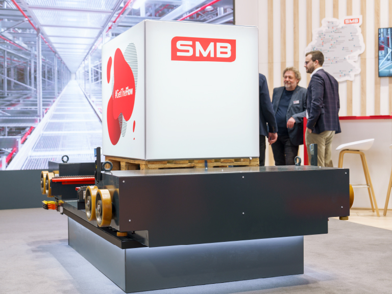 Ein automatisiertes Transportmodul der Firma SMB mit weiß-rotem Logo und dem Slogan „#GetTheFlow“, ausgestellt auf einem Messestand, im Hintergrund drei Personen im Gespräch.