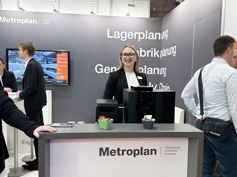Lächelnde Metroplan-Mitarbeiterin steht an einer modernen Messetheke mit Kaffeeautomat und Süßigkeiten, im Hintergrund Gespräche mit weiteren Besuchenden.