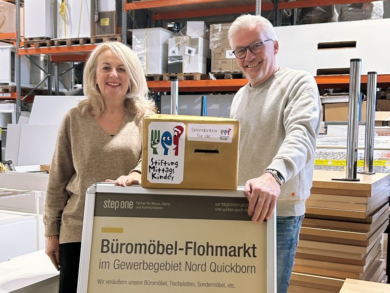 Eine Frau und ein Mann stehen in einem Lager neben einer Spendenbox für die Stiftung Mittagskinder. Vor ihnen ein Schild mit der Aufschrift „Büromöbel-Flohmarkt im Gewerbegebiet Nord Quickborn“.