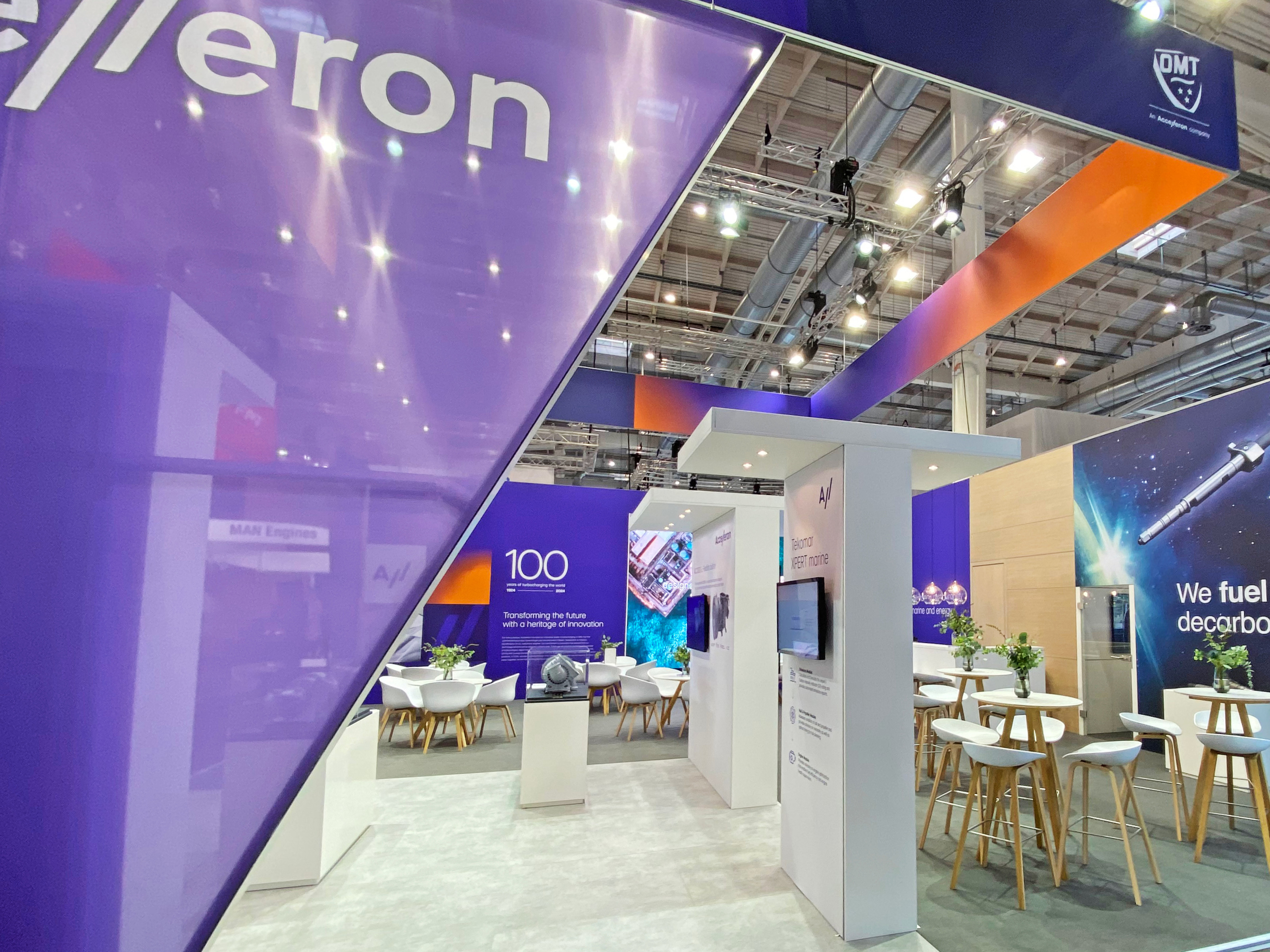 Messestand von Accelleron mit großem lila Markenelement im Vordergrund, modernen Sitzgruppen, Bildschirmen mit Präsentationen und stilvoller Beleuchtung in einer geräumigen Messehalle.