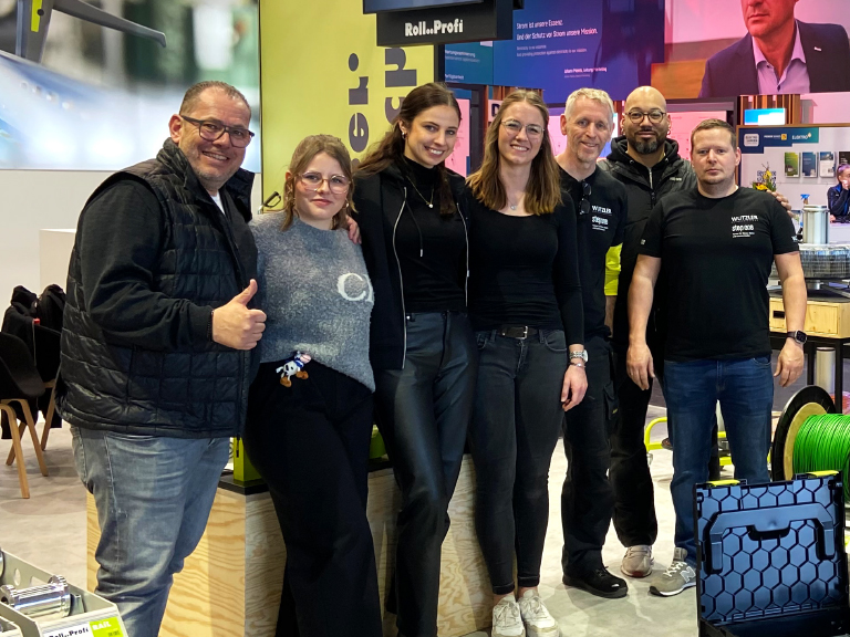 Sieben Teammitglieder von Wutzler posieren gut gelaunt auf dem Messestand vor dem Roll..Profi-Branding und zeigen Daumen hoch oder lächeln in die Kamera.