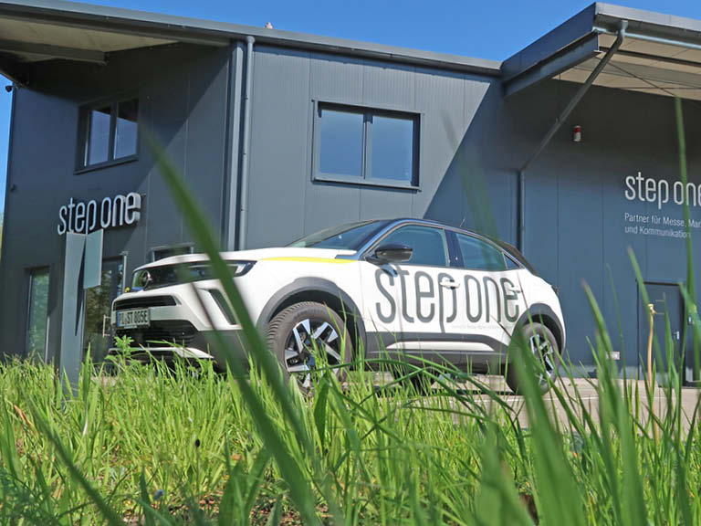 Elektroauto mit step one-Logo steht vor einem modernen Firmengebäude, fotografiert aus der Froschperspektive durch hohes grünes Gras bei sonnigem Wetter.