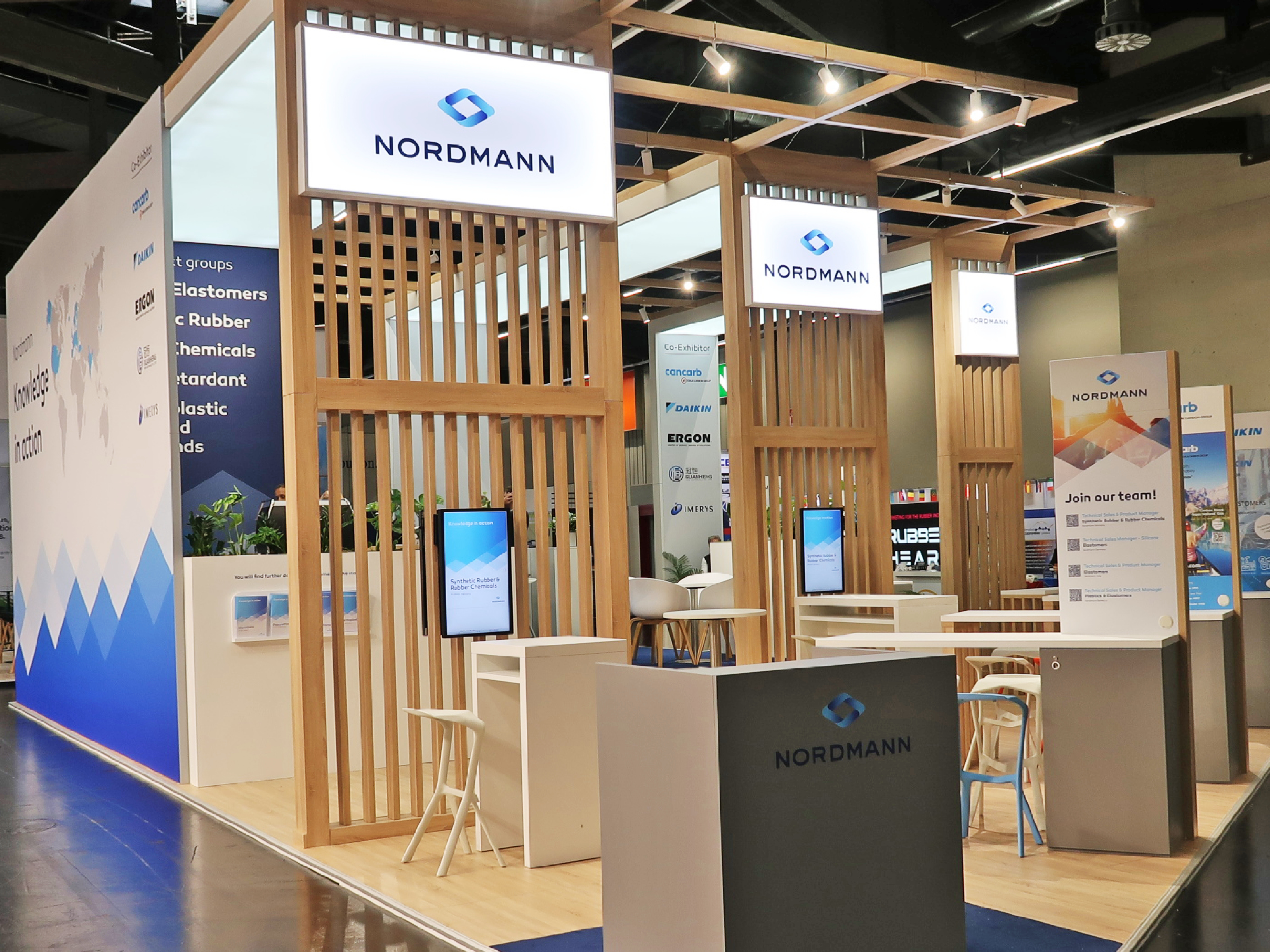 Nordmann-Messestand mit hohen Holzrahmen, beleuchteten Logos, Sitzmöglichkeiten, Informationsdisplays und einem modernen, strukturierten Aufbau in Natur- und Blautönen.