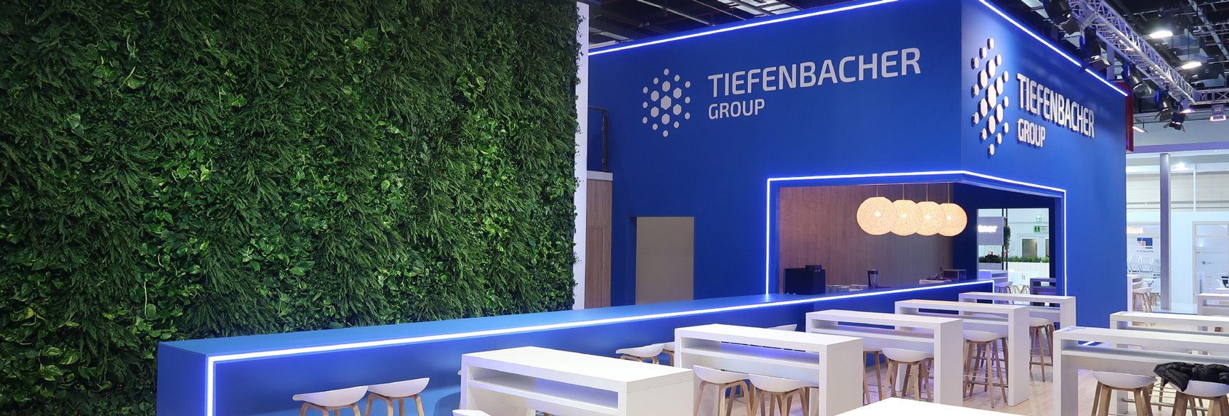 Ein Messestand mit einer großen grünen Pflanzenwand auf der linken Seite. Rechts befindet sich eine moderne, blau beleuchtete Theke mit hohen weißen Sitzhockern. Der Stand ist in Blau- und Holztönen gehalten, mit hängenden Lampen und einem offenen Bereich für Gespräche. Das Logo der „Tiefenbacher Group“ ist prominent auf der Wand zu sehen.