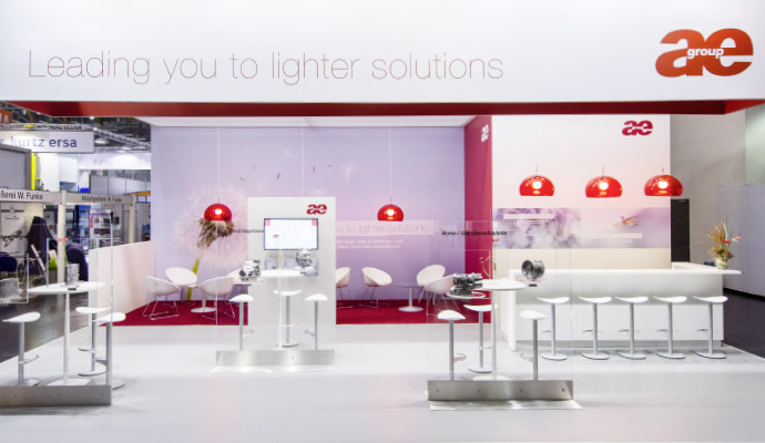 Ein offener Messestand mit einer weißen und roten Farbgestaltung. Ein großes weißes Banner mit der Aufschrift „Leading you to lighter solutions“ hängt oben. Mehrere weiße Barhocker stehen an einer Theke, und rote Hängeleuchten setzen Akzente. Im Hintergrund befinden sich Sitzgruppen mit weißen Sesseln und roten Teppichen.