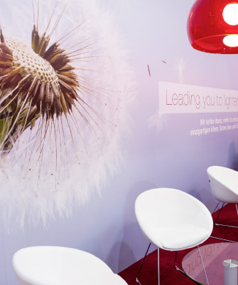 Eine gemütliche Sitzecke auf einem Messestand mit weißen Sesseln und Glas-Tischen auf rotem Teppich. An der Wand ist eine große Nahaufnahme einer Pusteblume mit dem Schriftzug „Leading you to lighter solutions“. Rote, halbtransparente Hängelampen sorgen für warmes Licht.