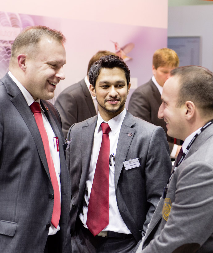 Drei Männer in Anzügen unterhalten sich lächelnd auf einer Messe. Sie tragen rote Krawatten und Namensschilder. Im Hintergrund sind weitere Personen, ein Messestand mit roten Lampen und eine dekorierte Bar zu sehen.