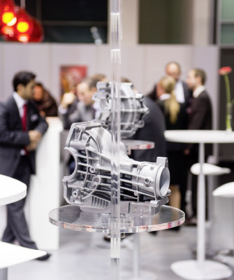 Ein metallisches Maschinenteil ist auf einer transparenten Acrylplatte befestigt, sodass es gespiegelt erscheint. Im Hintergrund sind mehrere Personen in Anzügen in geschäftlicher Unterhaltung auf einer Messe zu sehen.