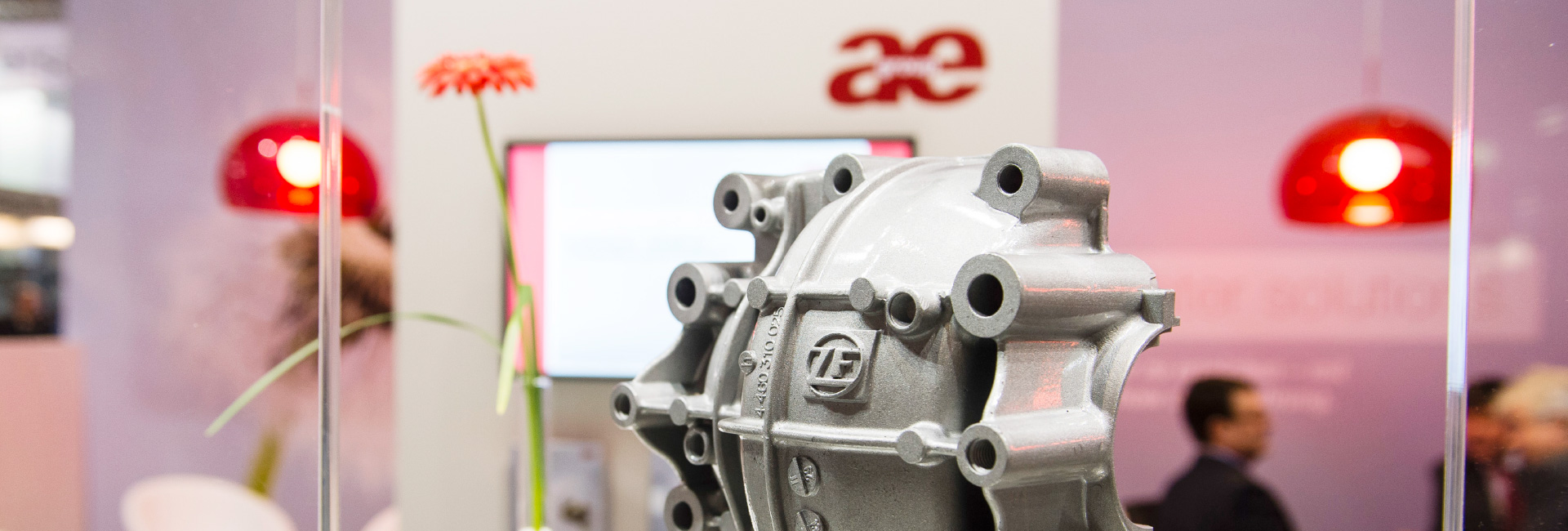 Nahaufnahme eines silbernen Metallbauteils mit dem Logo von ZF, präsentiert auf einer Messe. Im Hintergrund sind verschwommene Besucher, rote Hängelampen und ein Bildschirm mit Präsentationsinhalten zu sehen.
