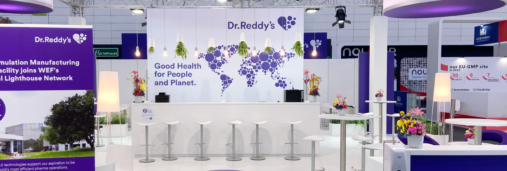 Ein großer Messestand von Dr. Reddy’s mit einer weißen Theke und mehreren Hockern davor. Die Rückwand zeigt das Logo des Unternehmens und eine Weltkarte aus violetten Kreisen mit dem Slogan „Good Health for People and Planet.“ Über der Theke hängen Lampen mit umgedrehten Pflanztöpfen. Rund um den Stand stehen kleine runde Tische mit bunten Blumensträußen. Im Hintergrund sind weitere Messestände mit Informationsmaterial zu pharmazeutischen Themen zu sehen.