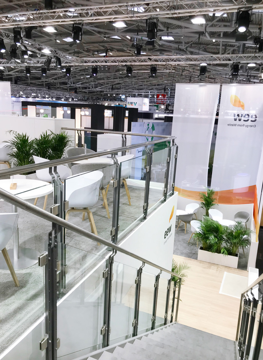 Ein zweistöckiger Messestand von EEW (Energy from Waste) mit einer Treppe, die zu einer oberen Lounge mit Glasgeländer führt. Dort stehen runde weiße Tische mit weißen und grauen Stühlen auf einem grauen Teppichboden. Unten gibt es weitere Sitzbereiche mit weißen Stühlen, runden Tischen und mehreren grünen Pflanzen in weißen Pflanzgefäßen. Große, transparente Banner mit dem EEW-Logo hängen an den Seiten. Im Hintergrund ist eine weitläufige Messehalle mit vielen Ständen und einer industriellen Beleuchtung mit zahlreichen schwarzen Scheinwerfern zu sehen.