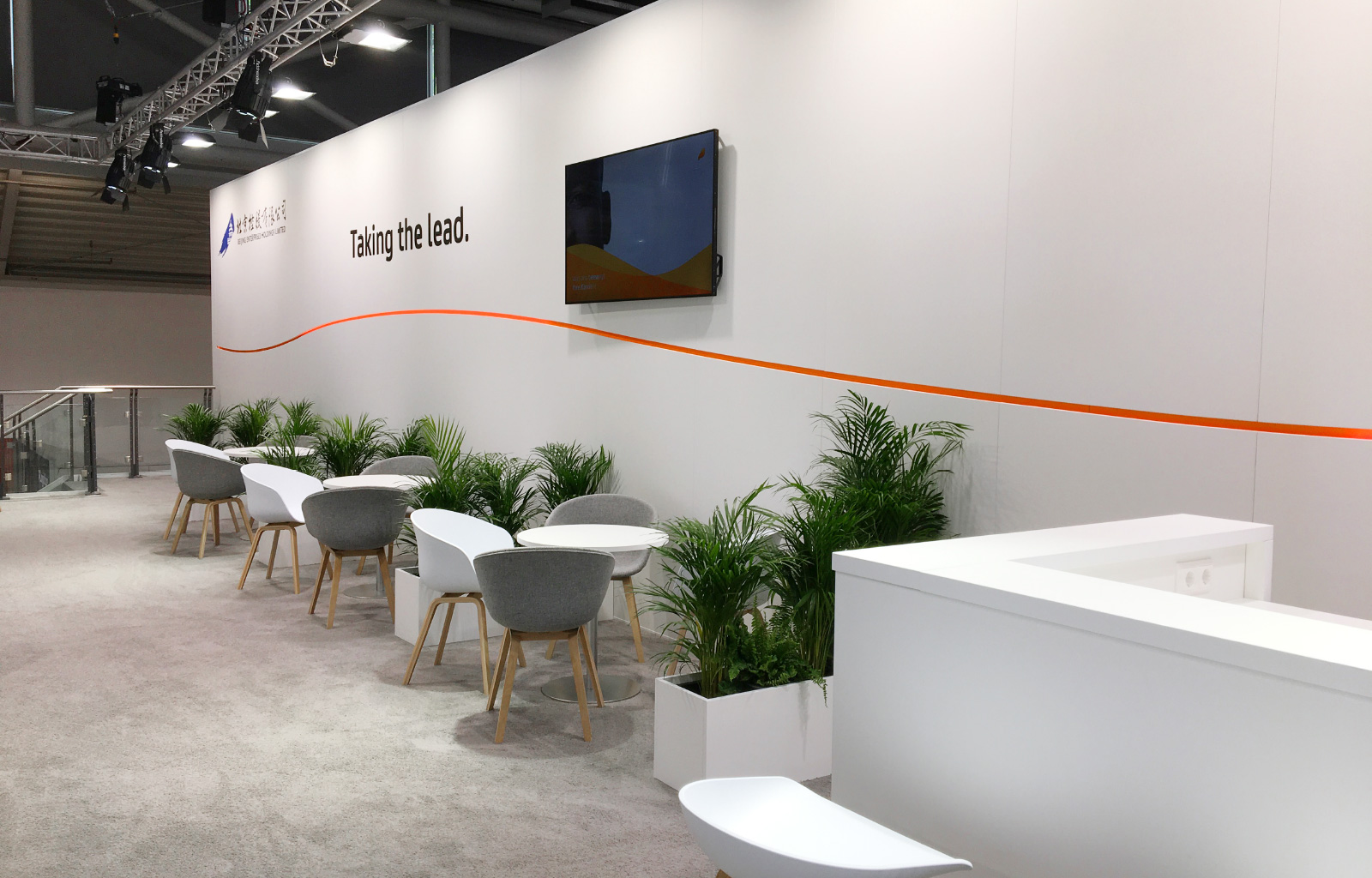 Ein stilvoll gestalteter Messestand mit einer weißen Rückwand, auf der der Slogan „Taking the Lead.“ und eine dezente orangefarbene Linie zu sehen sind. Davor befinden sich runde weiße Tische mit einer Mischung aus weißen und grauen Stühlen mit Holzbeinen. Mehrere grüne Pflanzen in weißen rechteckigen Pflanzgefäßen lockern das Design auf. Rechts steht ein moderner, weißer Empfangstresen mit integrierten Steckdosen. An der Wand hängt ein Bildschirm. Der Boden ist mit einem hellgrauen Teppich ausgelegt. Im Hintergrund ist ein Glasgeländer zu sehen, das auf eine untere Etage hinweist.