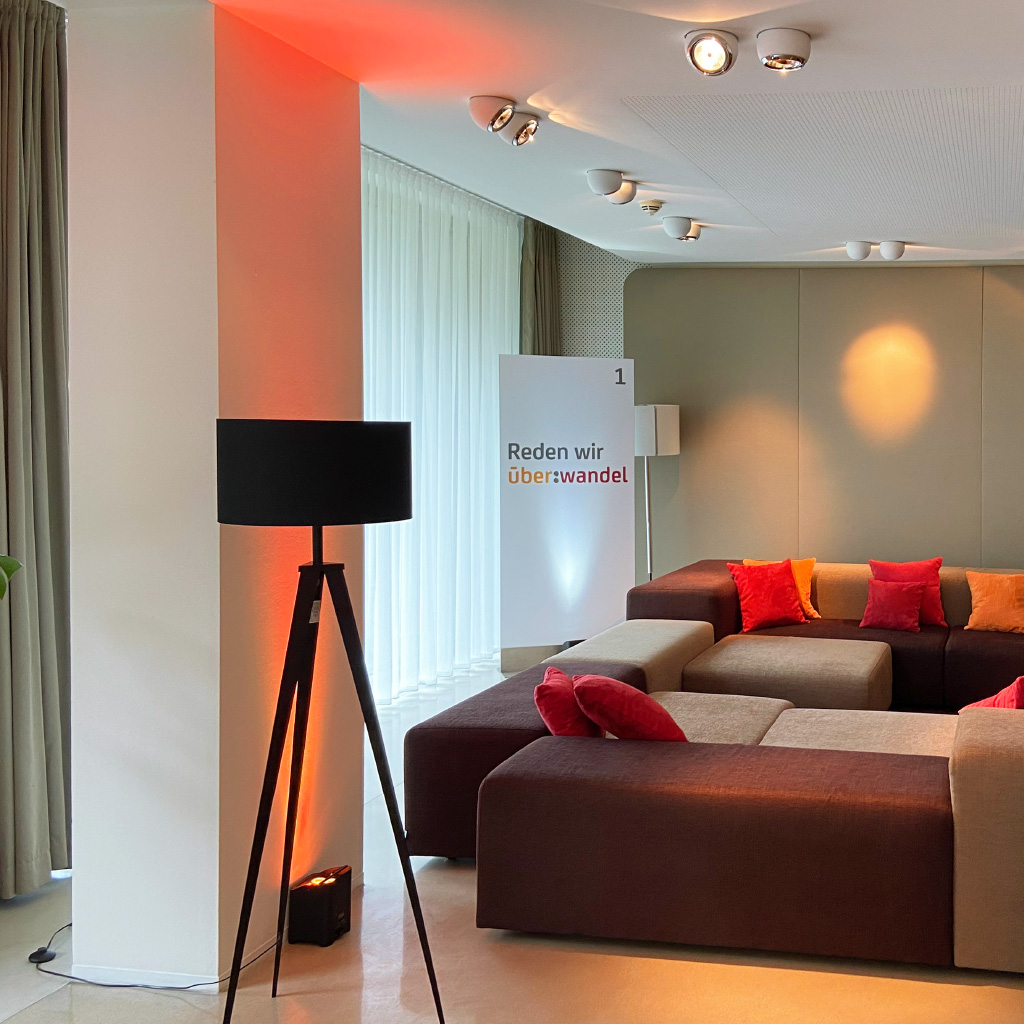 Ein stilvoll eingerichteter Loungebereich mit einer großen, modularen Sitzlandschaft in Brauntönen, dekoriert mit roten und orangefarbenen Kissen. Eine dreibeinige schwarze Stehlampe steht vor einer Wand mit indirekter, warm-oranger Beleuchtung. Im Hintergrund ist ein beleuchtetes Banner mit dem Schriftzug „Reden wir über: wandel“ zu sehen. Der Raum hat helle Vorhänge vor großen Fenstern, die Tageslicht hereinlassen, sowie eine moderne Deckenbeleuchtung.