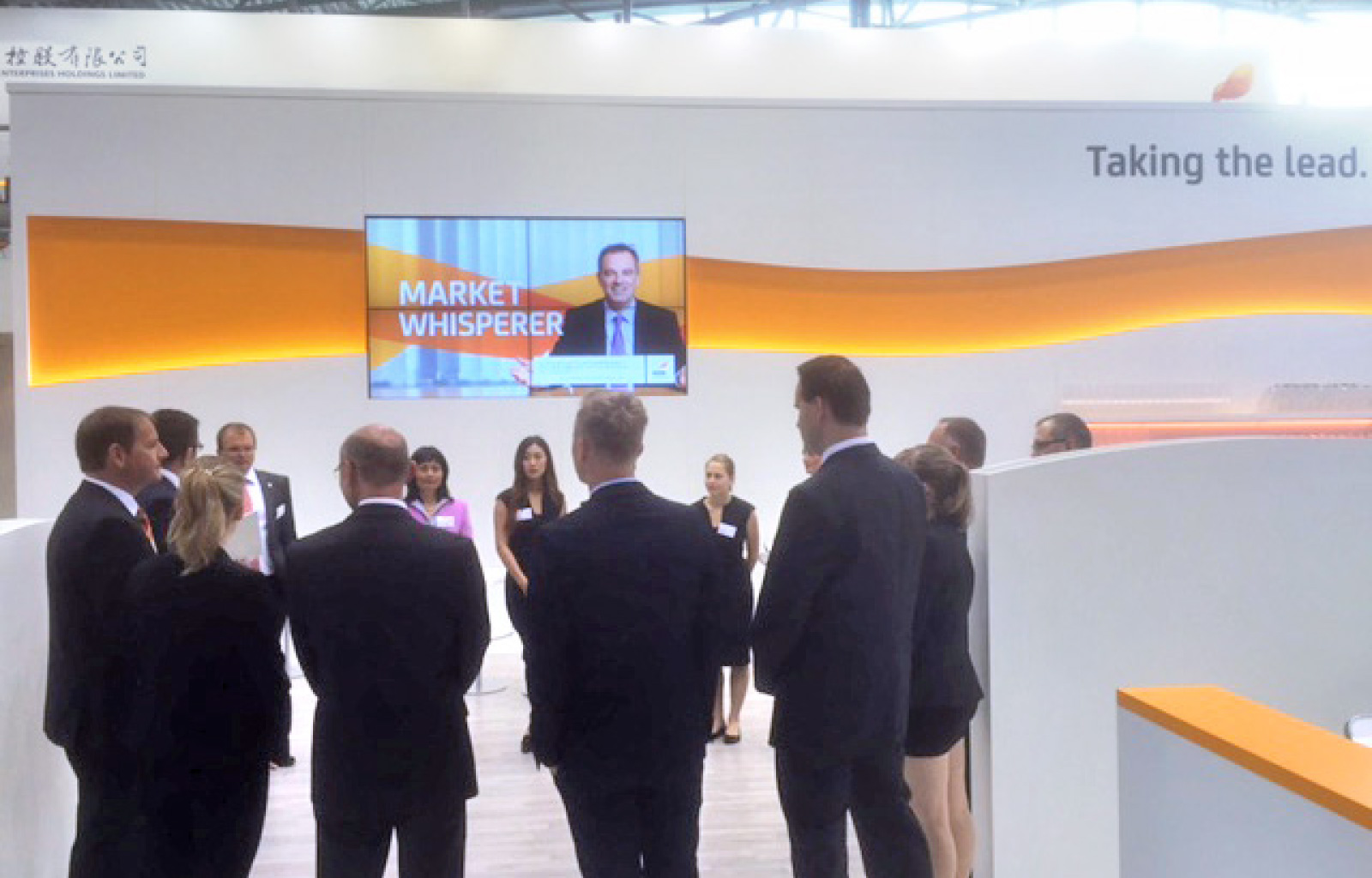 Mehrere Personen in Business-Kleidung stehen in einem Halbkreis an einem Messestand von EEW. Sie scheinen einem Briefing oder einer Präsentation zuzuhören. Der Stand ist modern gestaltet mit weißen Wänden und einer geschwungenen orangen Linie als Designelement. Im Hintergrund zeigt ein großer Bildschirm einen Sprecher mit der Aufschrift „Market Whisperer“. Der Slogan „Taking the lead.“ ist an der Wand zu sehen.