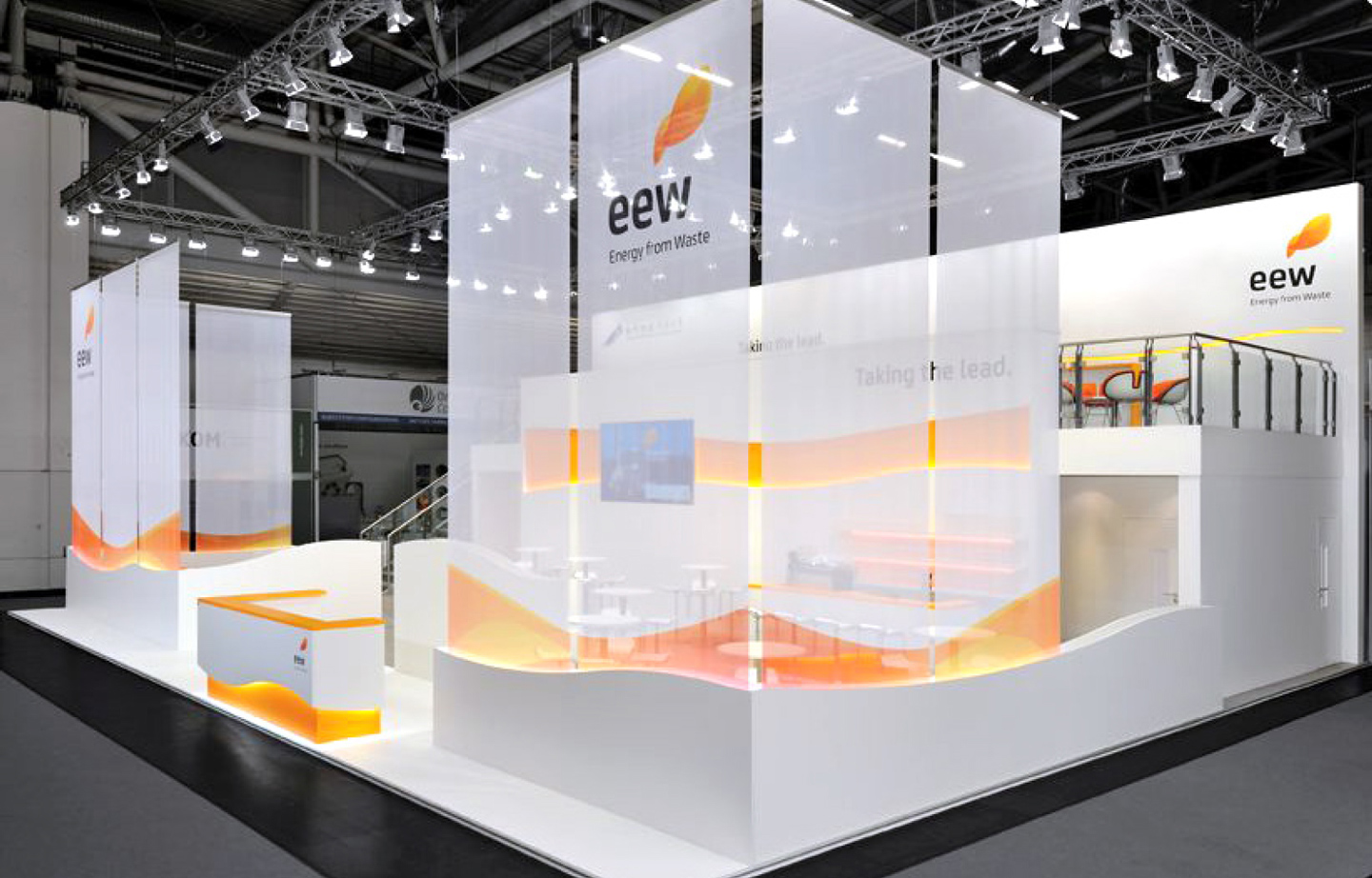 Ein großzügiger, moderner Messestand von EEW mit hohen weißen Wänden und transparenten Stoffbahnen, die den Stand umgeben. Das Design ist minimalistisch, mit geschwungenen weißen Elementen und orangefarbenen Akzenten. In der Mitte des Standes befindet sich eine offene Lounge mit weißen Tischen und Sitzgelegenheiten. Eine Treppe führt zu einer erhöhten Besprechungsebene mit Glasgeländer und orangen Stühlen. Beleuchtung und große Bildschirme ergänzen das moderne Design. Der Slogan „Taking the lead.“ ist an der Wand sichtbar.