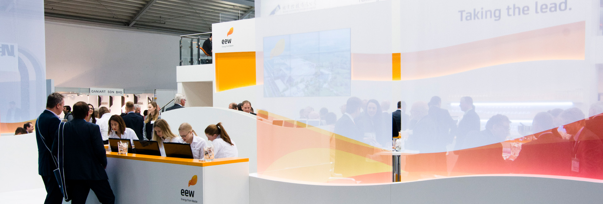 Ein lebendiger Messestand von EEW (Energy from Waste) mit einem geschwungenen, offenen Design. Im Vordergrund steht ein Empfangstresen mit mehreren Mitarbeiterinnen in weißen Hemden, die Besucher betreuen. Im Hintergrund ist eine Lounge mit Gästen an Tischen zu sehen. Große transparente Wände mit orangefarbenen Akzenten trennen die Bereiche. Auf einem Bildschirm werden Unternehmensinformationen angezeigt, während an der Wand der Slogan „Taking the lead.“ steht. Oberhalb des Standes befindet sich eine kleine, erhöhte Besprechungsebene mit Glasgeländer.