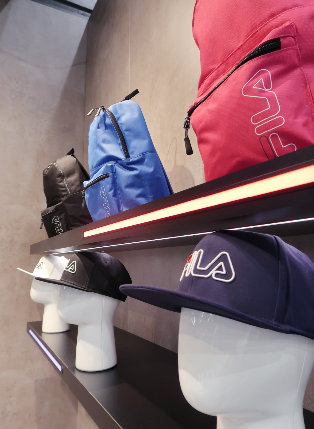 Ein Wandregal mit schwarzen Ablagen, auf denen Rucksäcke und Caps der Marke FILA ausgestellt sind. Die Rucksäcke in Rot, Blau und Schwarz stehen auf der oberen Ablage, während die Caps auf weißen Schaufensterköpfen auf der unteren Ablage platziert sind. Die Regale sind mit schmalen LED-Streifen in Rot und Weiß beleuchtet. Der Hintergrund besteht aus einer Betonoptik-Wand, die einen minimalistischen und modernen Look schafft. Im Vordergrund ist eine blaue Cap mit FILA-Logo deutlich sichtbar.