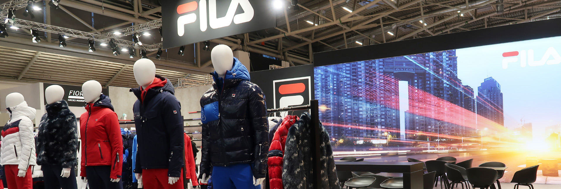 Ein FILA-Messestand mit Schaufensterpuppen, die Winterjacken und warme Sportbekleidung präsentieren. Die Kleidung ist in Blau-, Rot- und Weißtönen gehalten. Im Hintergrund sind ein großes FILA-Logo, ein Regal mit weiteren Produkten und eine LED-Wand mit einem dynamischen Stadtmotiv zu sehen. Rechts befindet sich ein Lounge-Bereich mit schwarzen Stühlen und Tischen. Die Szene ist modern beleuchtet und kombiniert urbane sowie sportliche Elemente.