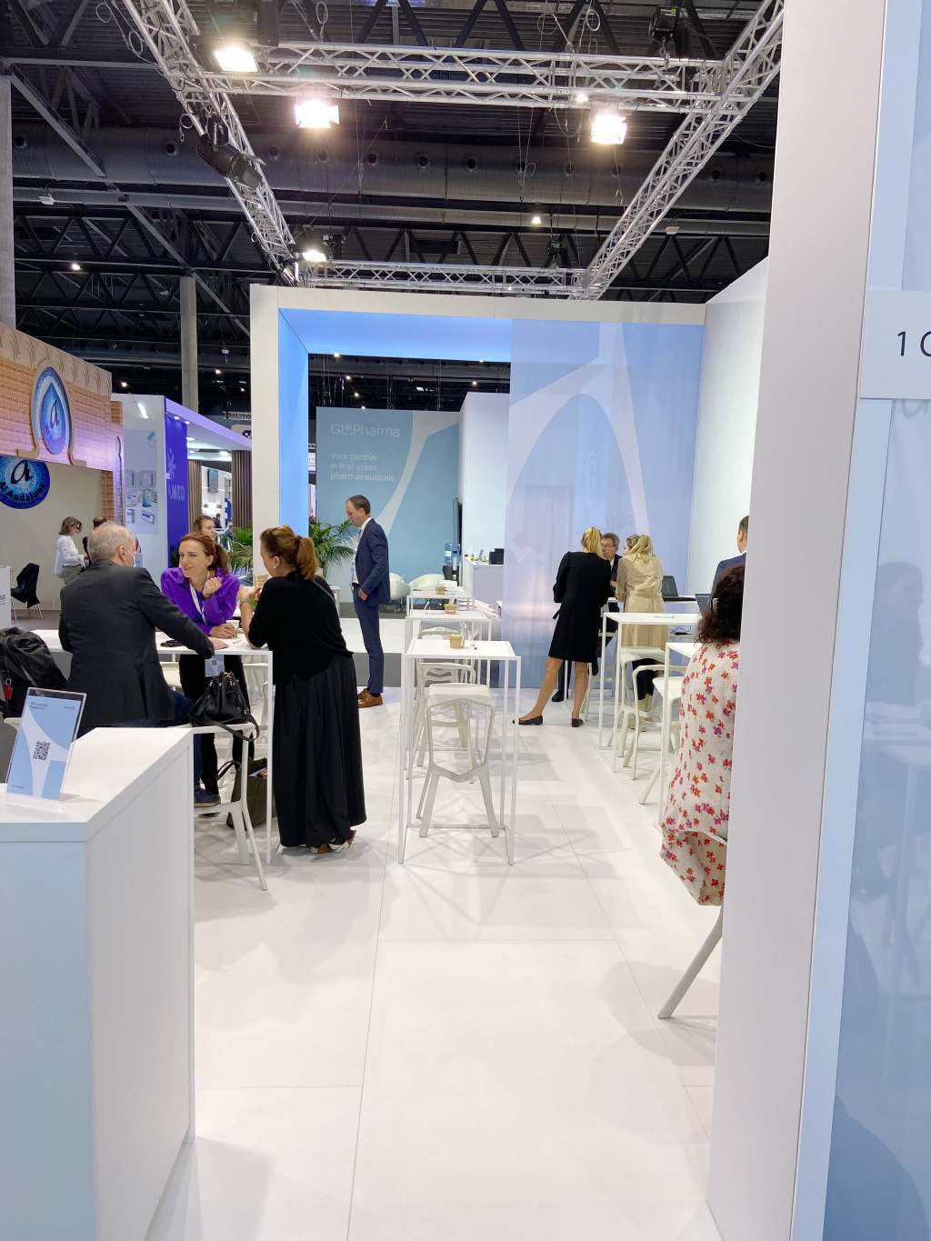 Messestand von GL Pharma mit weißen Stehtischen und Hockern. Mehrere Personen im Gespräch in einer offenen, modernen Umgebung.