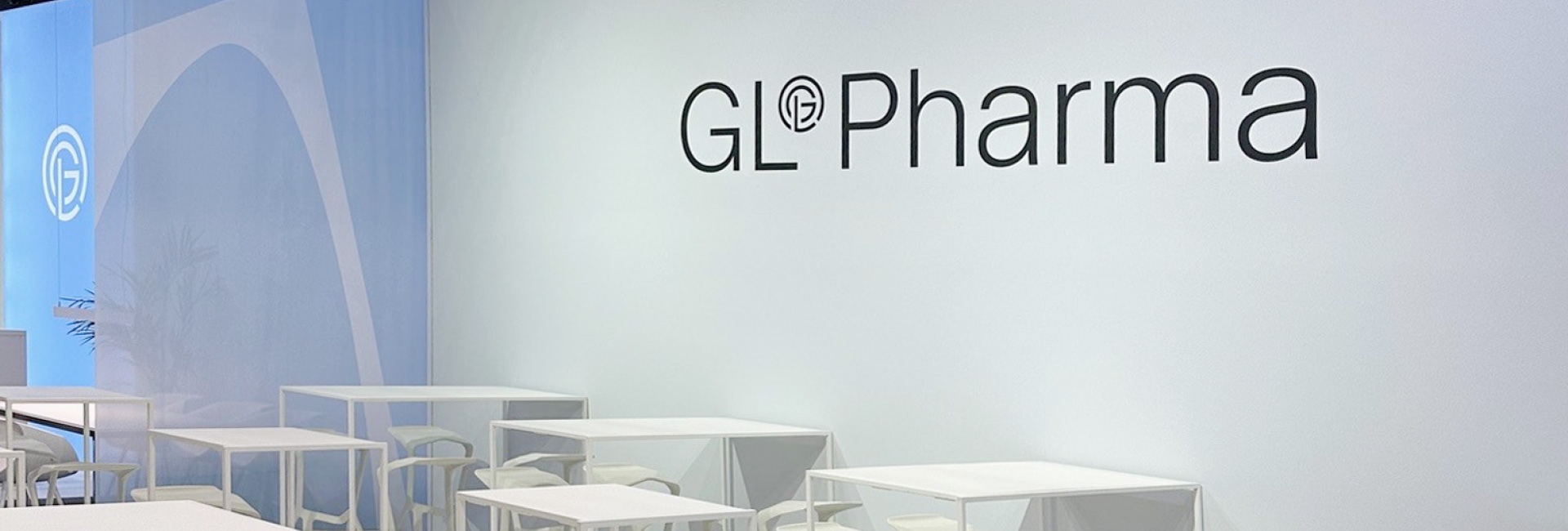 Nahaufnahme des GL Pharma Messestands mit weißer Wand, Logo und weißen Stehtischen mit Hockern in einem modernen Design.