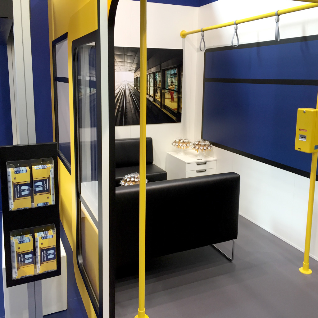 Messestand mit U-Bahn-Design, inklusive schwarzen Sitzbänken, Haltestangen und Ticketautomaten. Ein Bild eines Bahnsteigs sowie Prospekthalter mit Infomaterial sind zu sehen.
