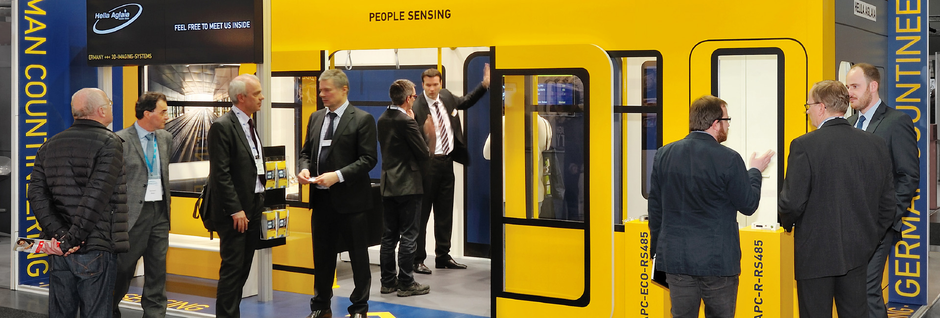 Messestand von Hella Aglaia in Gelb und Blau, gestaltet wie ein U-Bahn-Waggon. Mehrere Männer im Anzug führen Gespräche, während ein Bildschirm mit Informationen zu Fahrgastzählungstechnologien sichtbar ist. Die Standgestaltung umfasst Türen, Fenster und Haltegriffe, um eine realistische Bahnumgebung zu simulieren.