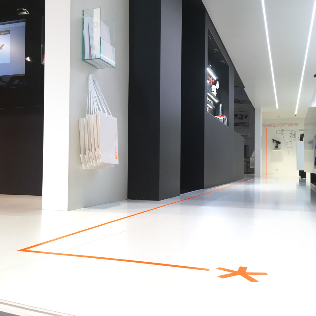 Innenansicht eines Messestands mit schwarzen und weißen Designelementen. Eine orangefarbene Bodenlinie mit einem Symbol am Anfang leitet die Besucher. An der Wand hängen weiße Tragetaschen mit orangefarbener Schrift sowie eine transparente Halterung mit Broschüren.