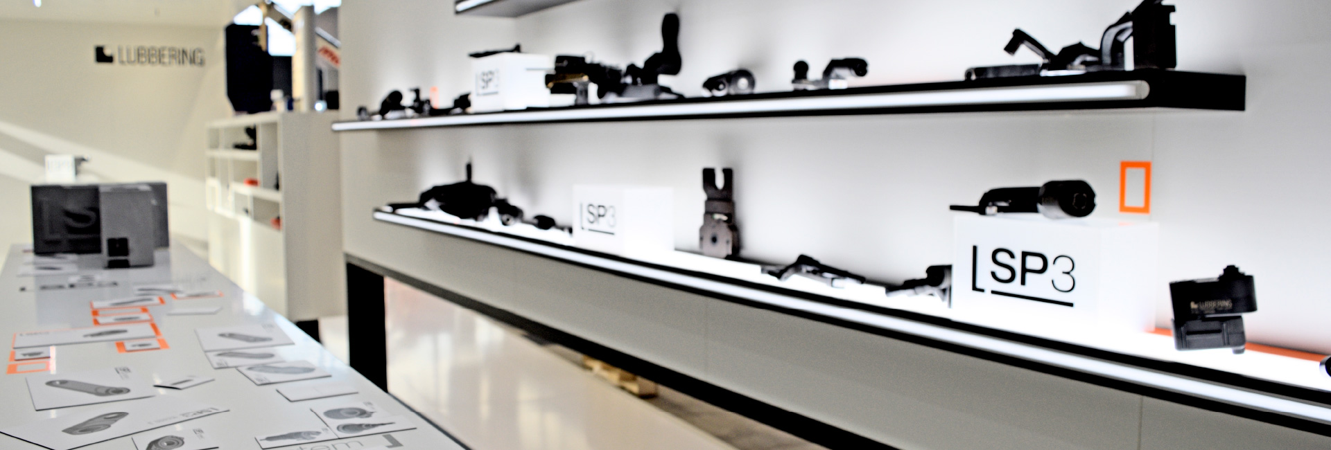 Ein modernes Ausstellungsregal mit schwarz-weißen Bauteilen, die auf beleuchteten Ebenen präsentiert werden. Im Vordergrund liegt eine Informationsfläche mit technischen Zeichnungen und Produktbeschreibungen.