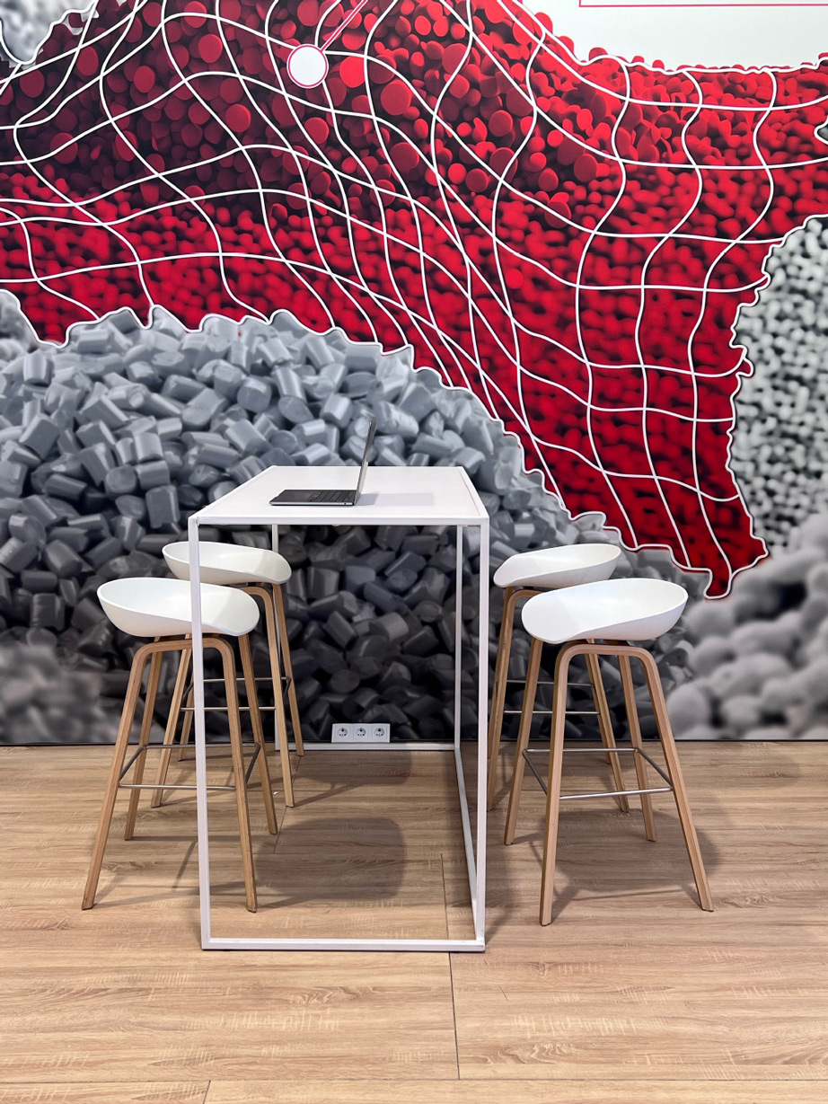 Ein schlichter weißer Tisch mit vier Barhockern mit Holzbeinen und weißen Sitzschalen steht vor einer auffälligen Wandgrafik mit roten und grauen Pellets, überzogen mit einem weißen Netz. Ein Laptop liegt auf dem Tisch, und eine Steckdose ist an der Wand sichtbar.