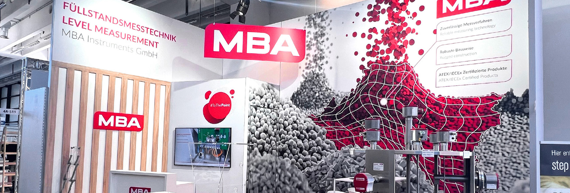 Ein Messestand mit der Aufschrift „Füllstandsmess­technik“ und dem MBA-Logo. Die Rückwand zeigt eine Grafik mit roten und grauen Pellets, überlagert von einem weißen Gitternetz. Ein technisches Exponat mit Sensoren ist auf dem Stand platziert. Ein Monitor zeigt digitale Inhalte.