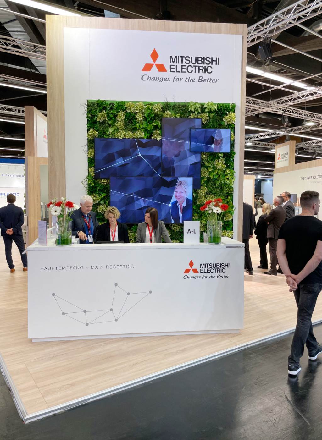 Ein Messestand von Mitsubishi Electric mit einem großen weißen Empfangstresen, auf dem das Unternehmenslogo und die Aufschrift „Hauptempfang – Main Reception“ zu sehen sind. Dahinter befinden sich drei Mitarbeiter mit roten Namensschildern. Die Rückwand ist mit einer grünen Pflanzenwand gestaltet und mit mehreren Monitoren bestückt, die digitale Inhalte anzeigen. Im Hintergrund sind Messebesucher und weitere Stände zu sehen.