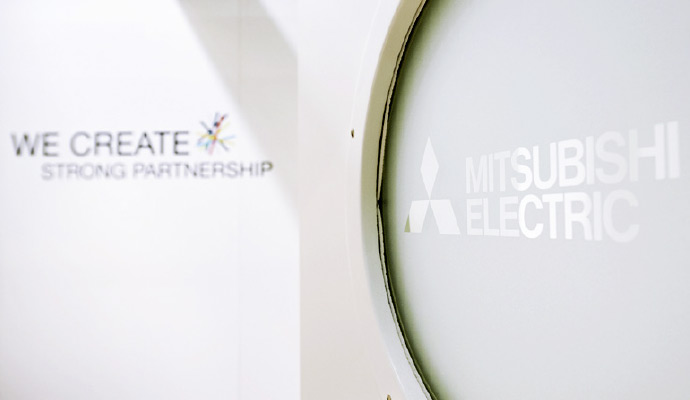 Eine Nahaufnahme eines Messebereichs von Mitsubishi Electric. Im Vordergrund ist eine runde, milchglasartige Oberfläche mit dem Firmenlogo „Mitsubishi Electric“ in heller Schrift zu sehen. Im Hintergrund steht auf einer weißen Wand der Slogan „We Create Strong Partnership“ mit einem farbigen grafischen Element.