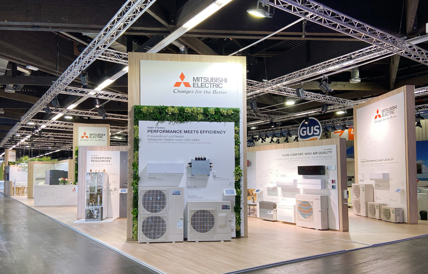 Ein Mitsubishi Electric Messestand auf einer Fachmesse mit modernen Heiz- und Kühlsystemen. Große Wände mit Produktpräsentationen zeigen Klimaanlagen, Wärmepumpen und Lüftungssysteme. Der Stand ist mit Holz- und Grünflächen gestaltet, während mehrere Infotafeln technische Details zu den Produkten liefern.