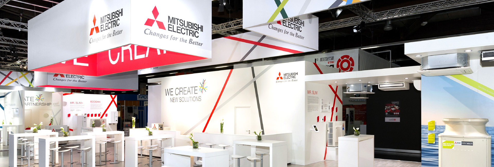 Ein großflächiger Mitsubishi Electric Messestand mit weißen, roten und grauen Designelementen. Der Stand zeigt verschiedene Heiz- und Kühlsysteme, darunter Wand- und Deckenklimageräte sowie Informationswände zu energieeffizienten Lösungen. Weiße Tische mit Barhockern und grünen Pflanzen sorgen für eine einladende Atmosphäre.