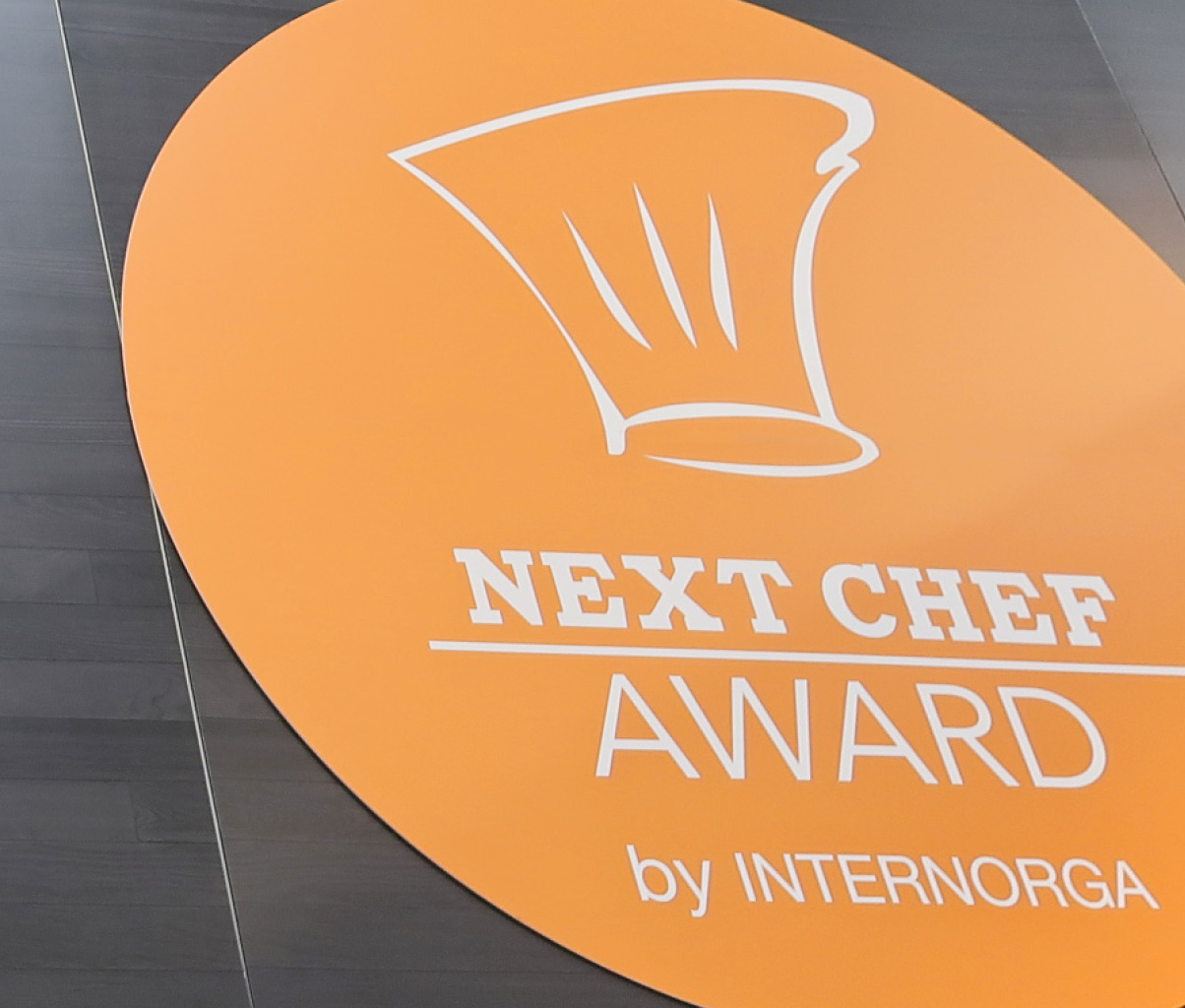 Großes, rundes, orangefarbenes Schild mit einer stilisierten Kochmütze und der Aufschrift „NEXT CHEF AWARD by INTERNORGA“. Das Schild befindet sich an einer Wand aus dunklem Holz.
