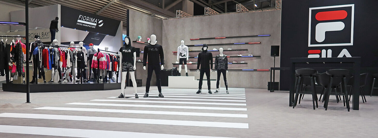 Ein FILA-Messestand mit mehreren Schaufensterpuppen, die Sportbekleidung präsentieren. Der Boden enthält ein Zebrastreifen-Design, das an eine städtische Straße erinnert. Im Hintergrund sind Schuhregale mit farbigen LED-Leisten und ein FILA-Logo auf einer schwarzen Wand sichtbar. Rechts gibt es einen Lounge-Bereich mit schwarzen Stühlen und Tischen. Links sind weitere Bekleidungsständer anderer Marken zu sehen. Der Stand kombiniert eine moderne, urbane Ästhetik mit sportlicher Eleganz.