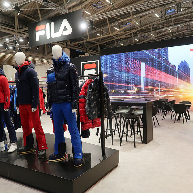 Ein FILA-Messestand mit Schaufensterpuppen, die Winterkleidung in Blau-, Rot- und Weißtönen tragen. Die Kleidung umfasst warme Jacken, Hosen und Winterstiefel. Im Hintergrund befindet sich eine große LED-Wand mit einer Stadtansicht und einem dynamischen Lichteffekt. Daneben gibt es einen Lounge-Bereich mit schwarzen Stühlen und Tischen sowie ein großes FILA-Logo über dem Stand. Die Szene ist modern beleuchtet und kombiniert Sport- und Lifestyle-Elemente.