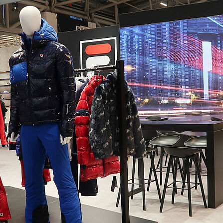Ein FILA-Messestand mit Schaufensterpuppen, die Winterkleidung in Blau-, Rot- und Weißtönen tragen. Die Kleidung umfasst warme Jacken, Hosen und Winterstiefel. Im Hintergrund befindet sich eine große LED-Wand mit einer Stadtansicht und einem dynamischen Lichteffekt. Daneben gibt es einen Lounge-Bereich mit schwarzen Stühlen und Tischen sowie ein großes FILA-Logo über dem Stand. Die Szene ist modern beleuchtet und kombiniert Sport- und Lifestyle-Elemente.