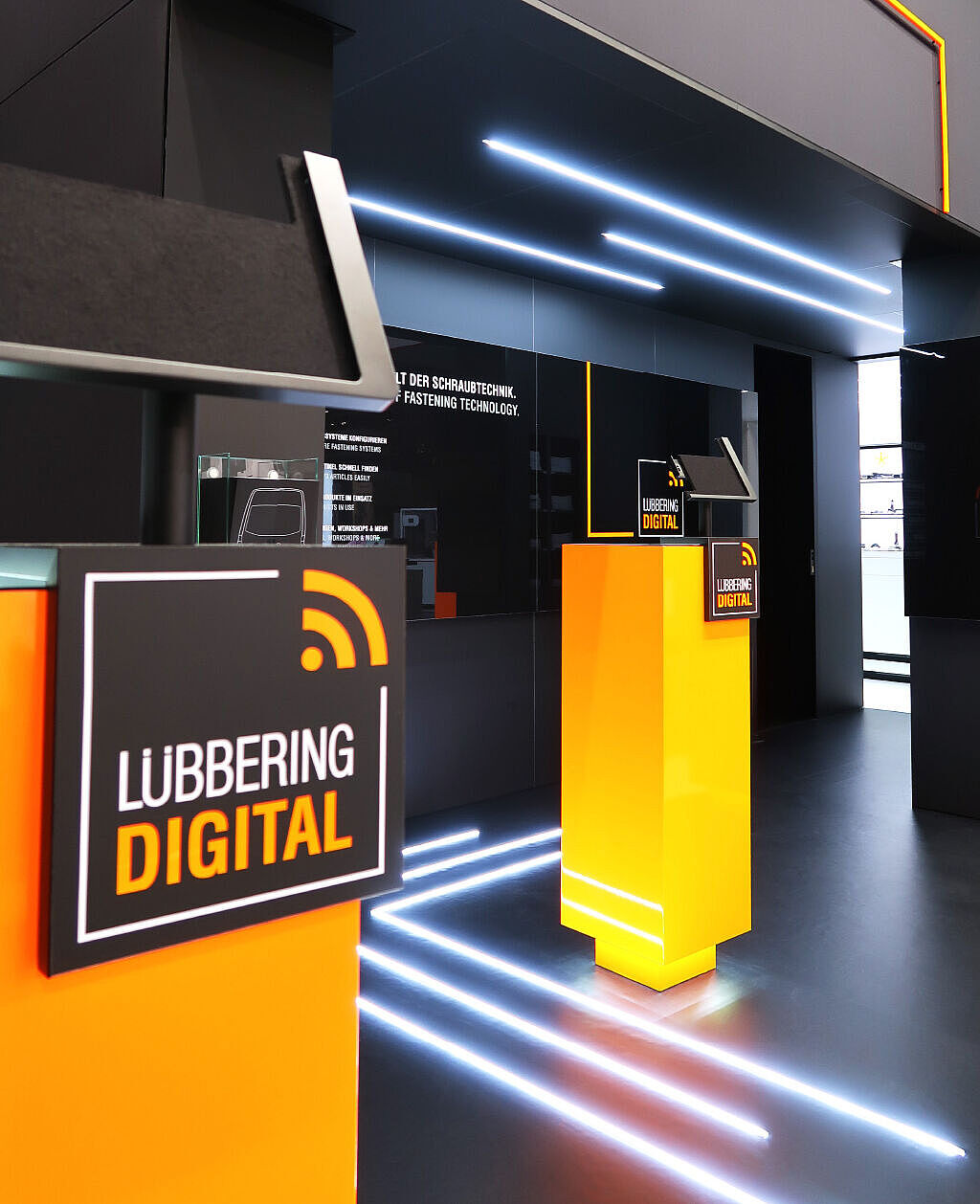 Messestand von LÜBBERING DIGITAL mit leuchtenden orangefarbenen Podesten und schwarzen Schildern mit weißer und orangefarbener Schrift. LED-Streifen verlaufen am Boden und an den Wänden, während ein moderner, digital inspirierter Look den Bereich prägt.